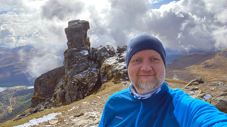 Gordon McLean takes a selfie up a Scottish mountain. There is an interesting rock formation and a bank of clouds in the background.