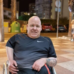 Gerry Dalrymple sits in his wheelchair in what appears to be an indoor space with parked cars visible in the background, wearing a dark Nike athletic shirt. He's smiling at the camera with his hands resting on his lap, positioned in front of what looks like a parking area or garage setting.