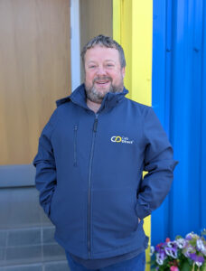 Stuart Sloan stands smiling with hands in his jacket pockets. He wears a navy blue softshell jacket branded with the “Cab Direct” logo in white and yellow. Behind him is a brightly coloured setting featuring a yellow door frame, blue wall panelling, and a pot of mixed flowers at the bottom right.
