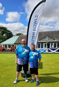 Stuart Sloan, on the right, stands with his teammate on a bowling green in front of a "British Bowls" flag. Both wear blue team Scotland shirts with abstract white designs and gold medals. Stuart is shorter, with dark hair and a beard, while his teammate is taller, bald, and has a black prosthetic leg. Behind them is a clubhouse decorated with two Scottish flags and people in red and green shirts.