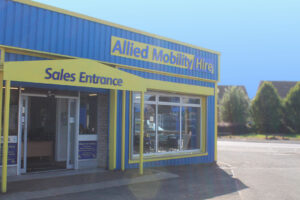 Exterior of a building with blue and yellow signage reading ‘Allied Mobility Hire.’ A large window is on the right, and an entrance on the left has a sign above it that says ‘Sales Entrance.