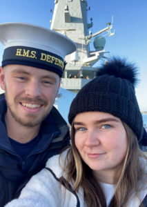 Selfie of Elisha Fitzpatrick and David on a ship. David is wearing a navy uniform with a hat that reads 'H.M.S DEFENDER.' Elisha is wearing a black knit hat with a pom-pom and a white jacket. The ship's structure is visible in the background.
