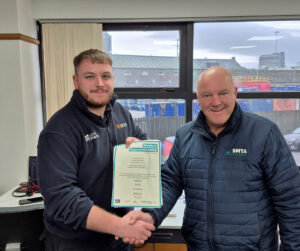 Aaron Rennie is shaking hands with another person indoors while holding a certificate titled “Modern Apprenticeship.” The certificate includes text: “Modern Apprenticeship This is to certify that Aaron Rennie has successfully completed a Modern Apprenticeship in Automotive Parts Operations at SCQF Level 6 Awarded by Institute of the Motor Industry Signed by Assessor and Internal Verifier.” Both individuals wear branded jackets, and a window behind them shows an industrial outdoor setting.