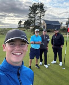 Four people standing on a golf green near a hole with a red flag, holding golf clubs. The person in the foreground wears a blue patterned jacket and a dark cap. Behind, one person wears a light blue striped polo shirt and navy shorts, another wears a dark top with black trousers, and the third wears a black jacket with white trainers. The background shows a modern stone building, trees, and a cloudy sky.