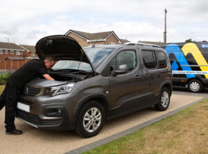 A mechanic in black workwear leans over the open bonnet of a grey Peugeot van parked on a residential drive. A branded service van with blue and yellow markings is parked beside it, with red‑brick houses and a cloudy sky in the background.