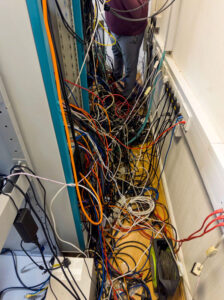 Cluttered server room with tangled cables in orange, red, and black running between racks and power strips along the wall, creating a chaotic and unorganised network environment.