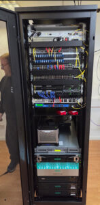 Tall server rack filled with networking equipment including switches, routers, patch panels, and storage units. Cables are neatly organised by colour—yellow, blue, and red—indicating a professional IT setup.