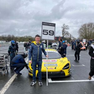 A person in a padded jacket stands on a wet starting grid holding a sign marked 59 before a yellow race car covered in sponsor logos including "DUCKHAMS." Crew members work around the vehicle under grey, rainy skies.