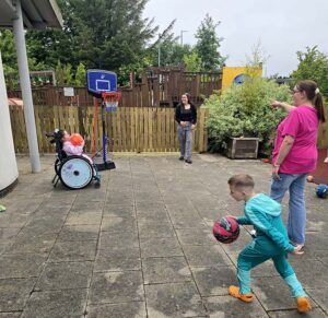 A child in a wheelchair shoots a basketball whilst another child runs with a ball. Two adults watch nearby. There is a wooden fence, playground equipment, and greenery in the background.