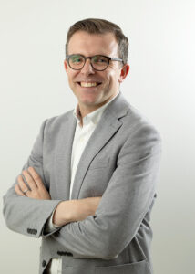 A man wearing glasses, a light grey blazer, and a white shirt stands with arms crossed, smiling at the camera against a plain white background.