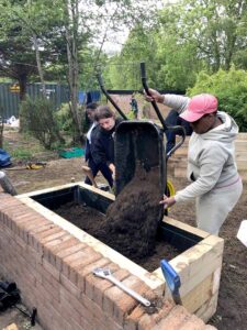 People tip soil from a wheelbarrow into a raised brick planter outdoors. One person grips the handles while others guide the load. Trees, tools and timber boards surround the planting area.