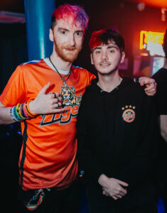 Ducky and Robert pose together for a photo in a nightclub. Ducky wears an orange Tigers sports top and Robert wears a Tigers hoodie.