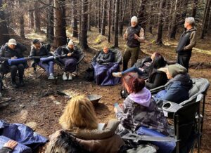 A group of people sit in a circle on camping chairs in a forest, listening to a person standing and speaking. The scene appears relaxed and informal, with sunlight filtering through the trees.