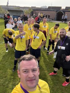 A group of football players in yellow shirts pose for a selfie on a grassy pitch, wearing medals, with spectators and buildings in the background. Everyone appears happy and relaxed after a match.