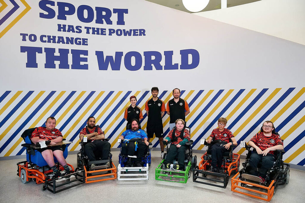 Eight people, some in powered wheelchairs and some standing, wear matching sports kits. They pose in front of a wall that reads, Sport has the power to change the world. Diagonal yellow and blue lines decorate the wall.