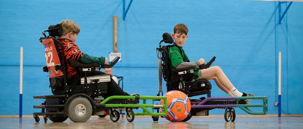 Two boys in powered wheelchairs play a game of powerchair football indoors, with a large orange football between them on a gym floor.