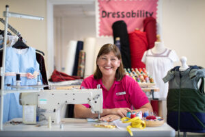 A woman in a pink shirt sits at a sewing machine, smiling, surrounded by fabric, thread, and clothing. A Dressability sign hangs behind her, with dresses and rolls of fabric visible in the background.