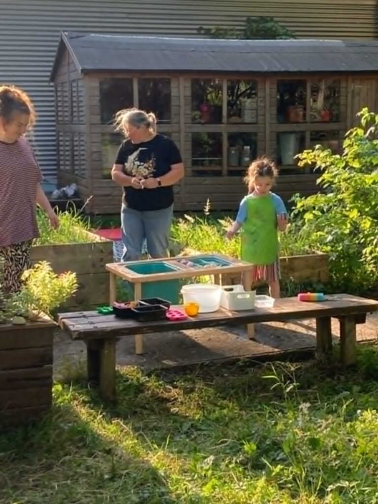 Three people, including a child in a green apron, stand near a wooden table with bowls and tools outside in a garden area. A wooden shed with windows is visible in the background.