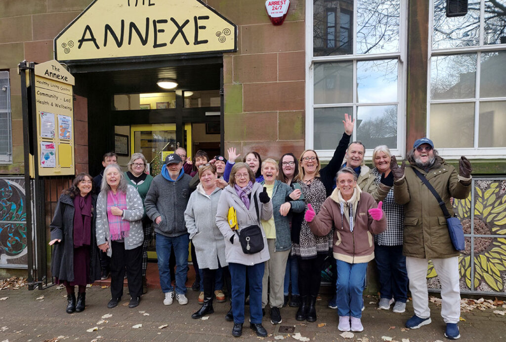 A diverse group of adults smiling and posing for a photo outside the entrance to The Annexe community centre.
