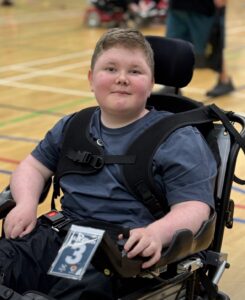 A young boy sitting in a powered wheelchair in a gymnasium, with the number 3 displayed on his panel.
