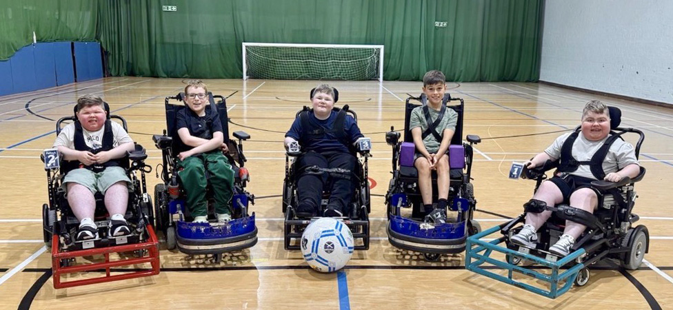 Five children in powered wheelchairs with sport guards lined up on a gymnasium floor with a power football in the centre.