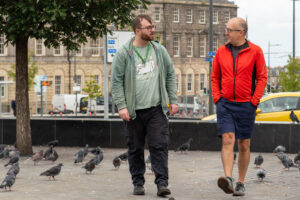 Two men walking and talking in a city square filled with pigeons.