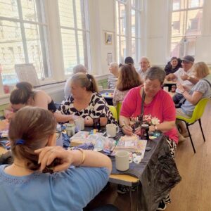 A group of people sitting at tables in a bright, sunlit room, painting small stones together.